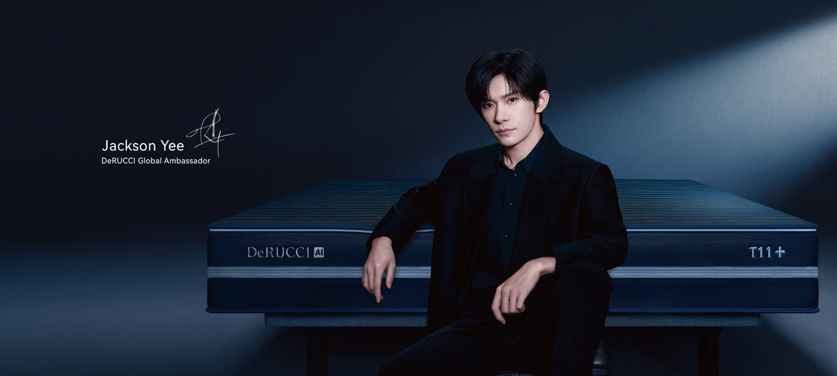 Jackson yee in a black suit sits on a dark blue de'rucci t11+ mattress in a dimly lit studio setting
